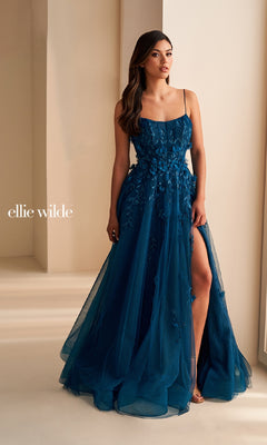 A woman in the "Ellie Wilde EW36222" long blue prom dress, featuring floral details, a high slit, and lace applique, stands indoors by a cream-colored wall. The text "ellie wilde" appears on the left.