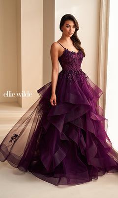 A woman stands indoors in the Ellie Wilde EW36225, a deep purple long prom dress with floral embroidery, spaghetti straps, and a voluminous tulle skirt. "ellie wilde" appears on the image.