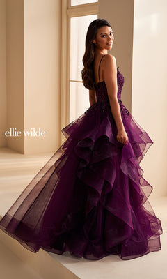 A woman in a purple, tiered tulle ball gown with spaghetti straps and floral accents stands by a window, glancing over her shoulder. She wears the stunning Ellie Wilde EW36225 long prom dress.
