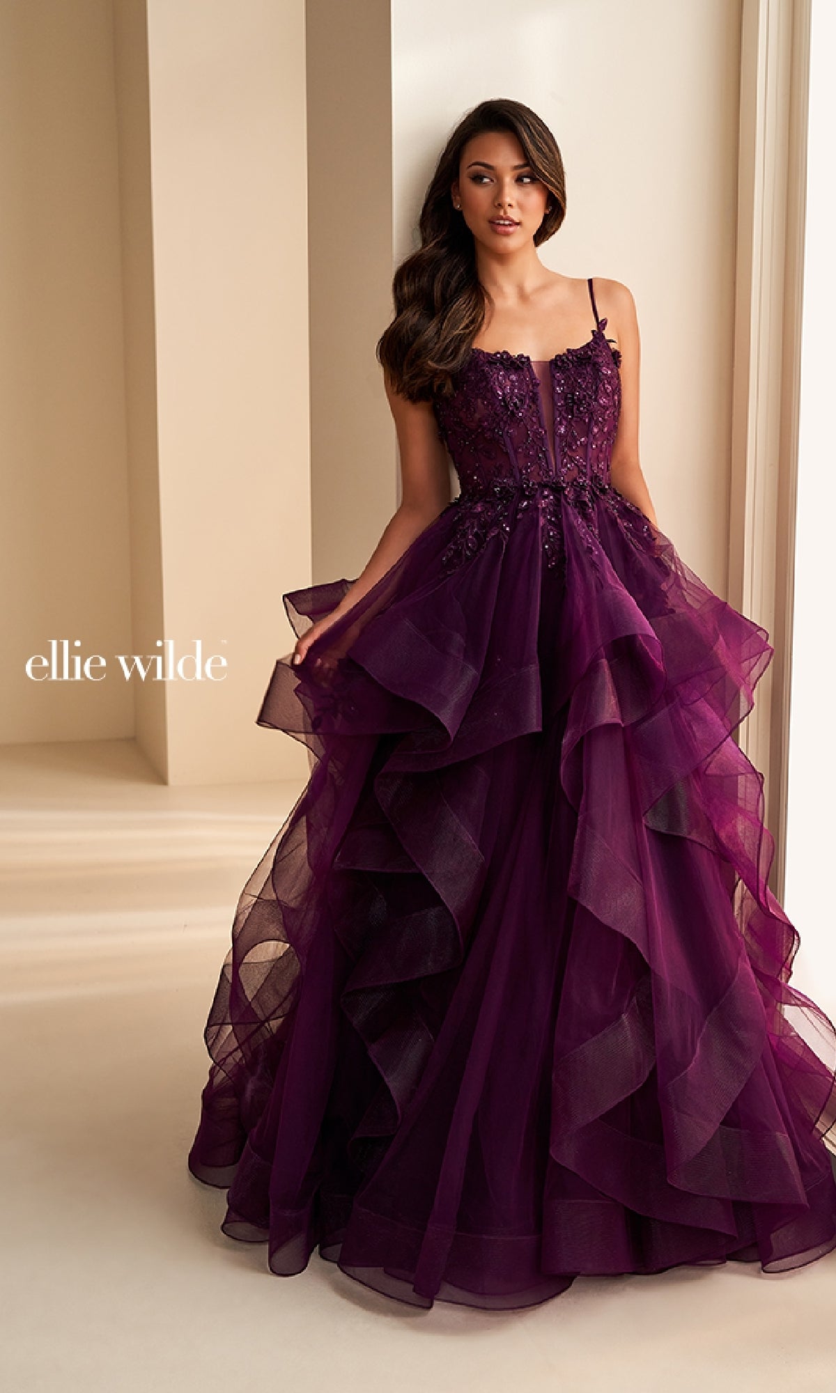A woman stands indoors near a cream-colored wall wearing the Ellie Wilde EW36225 long prom dress—a deep purple, strapless ball gown featuring tiered ruffles and floral embellishments.