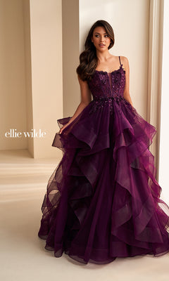A woman stands indoors near a cream-colored wall wearing the Ellie Wilde EW36225 long prom dress—a deep purple, strapless ball gown featuring tiered ruffles and floral embellishments.