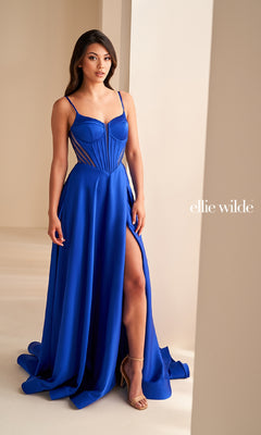 A woman stands indoors by a light wall wearing the Ellie Wilde EW36227 long royal blue satin A-line prom dress with spaghetti straps, corset bodice, thigh-high slit, and sheer paneling. "ellie wilde" is visible on the right.