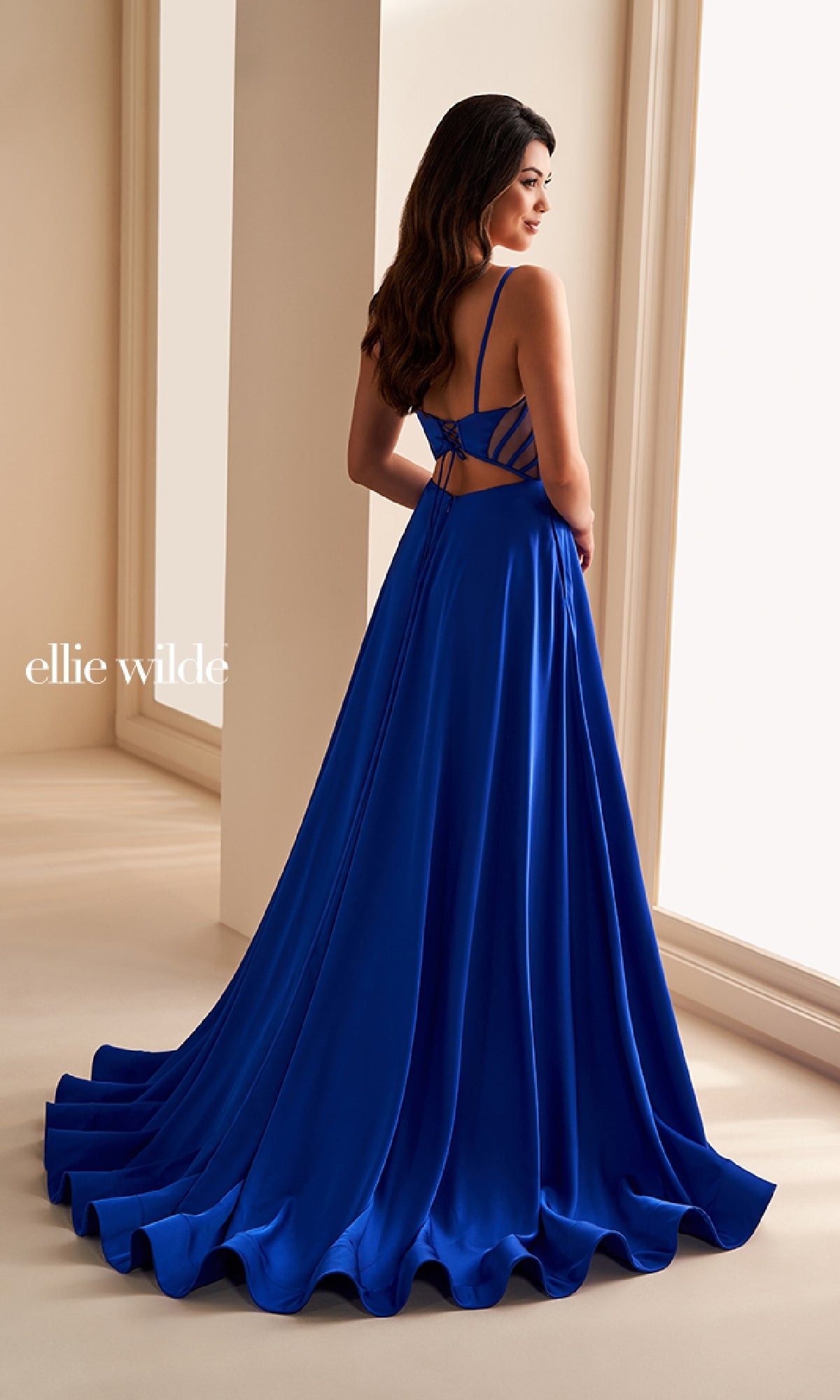 A woman stands near large windows, facing away in the Ellie Wilde EW36227—a royal blue, floor-length satin a-line prom dress featuring a strappy open back and wide, smooth folds flowing elegantly to the floor.