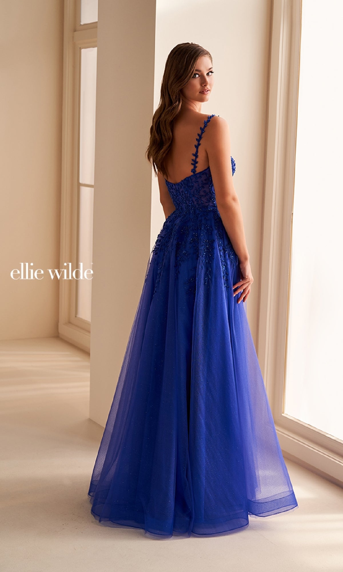 A woman stands indoors wearing the Ellie Wilde EW36228 long prom dress, a royal blue gown with lace appliques, a corset bodice, and sheer overlay, as she looks over her shoulder.