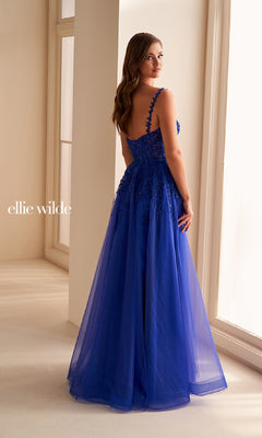 A woman stands indoors wearing the Ellie Wilde EW36228 long prom dress, a royal blue gown with lace appliques, a corset bodice, and sheer overlay, as she looks over her shoulder.