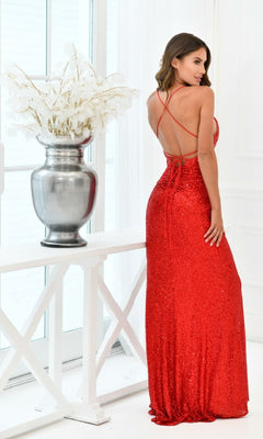 A woman stands indoors beside a large silver vase with white branches, wearing the Eclipse by Velvi—an open-back long sequin prom dress in elegant red.