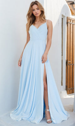 A woman in the Simple V-Neck Long A-Line Prom Dress 477—a light blue, floor-length gown with a thigh-high slit—stands near a white wall and wooden door, paired with silver high-heeled sandals.