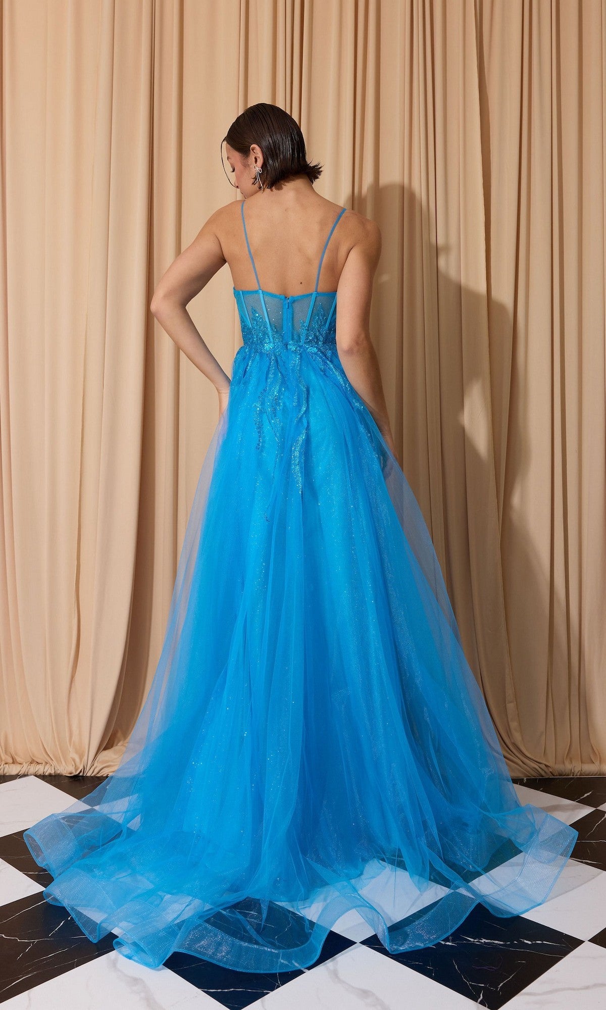 A woman faces away from the camera, wearing the Overskirt Long Sequin Prom Dress: Eureka EK112—a bright blue, floor-length tulle gown with a corset-style, sequin bodice—standing before beige curtains on a black-and-white floor.