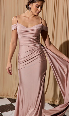 A woman in the Eureka EK117 off-shoulder long formal dress in blush pink, featuring draped fabric, stands before beige curtains on a checkered floor—a stunning choice for any special occasion.
