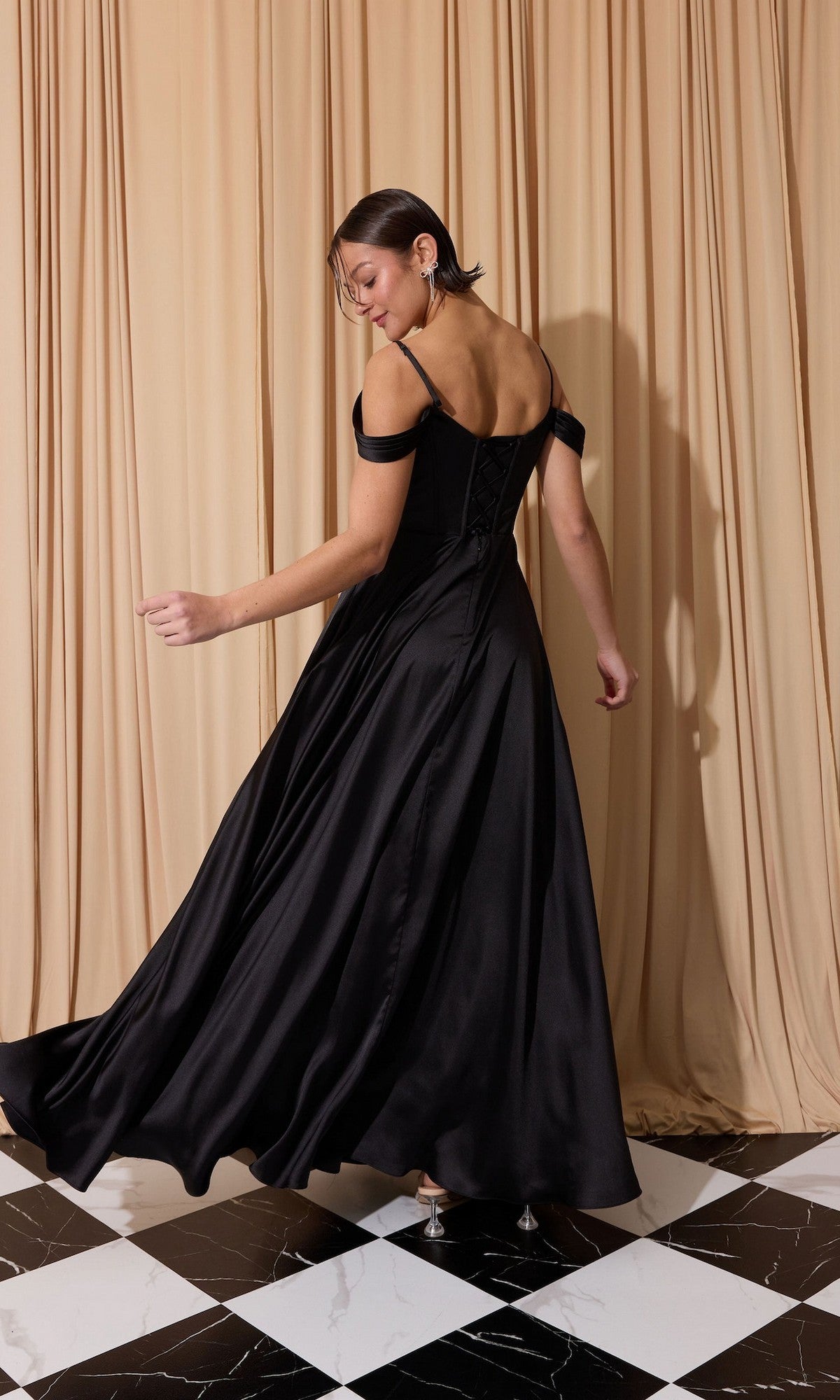 A person wearing the Eureka EK176 long prom dress poses on a black and white checkered floor in front of beige curtains.