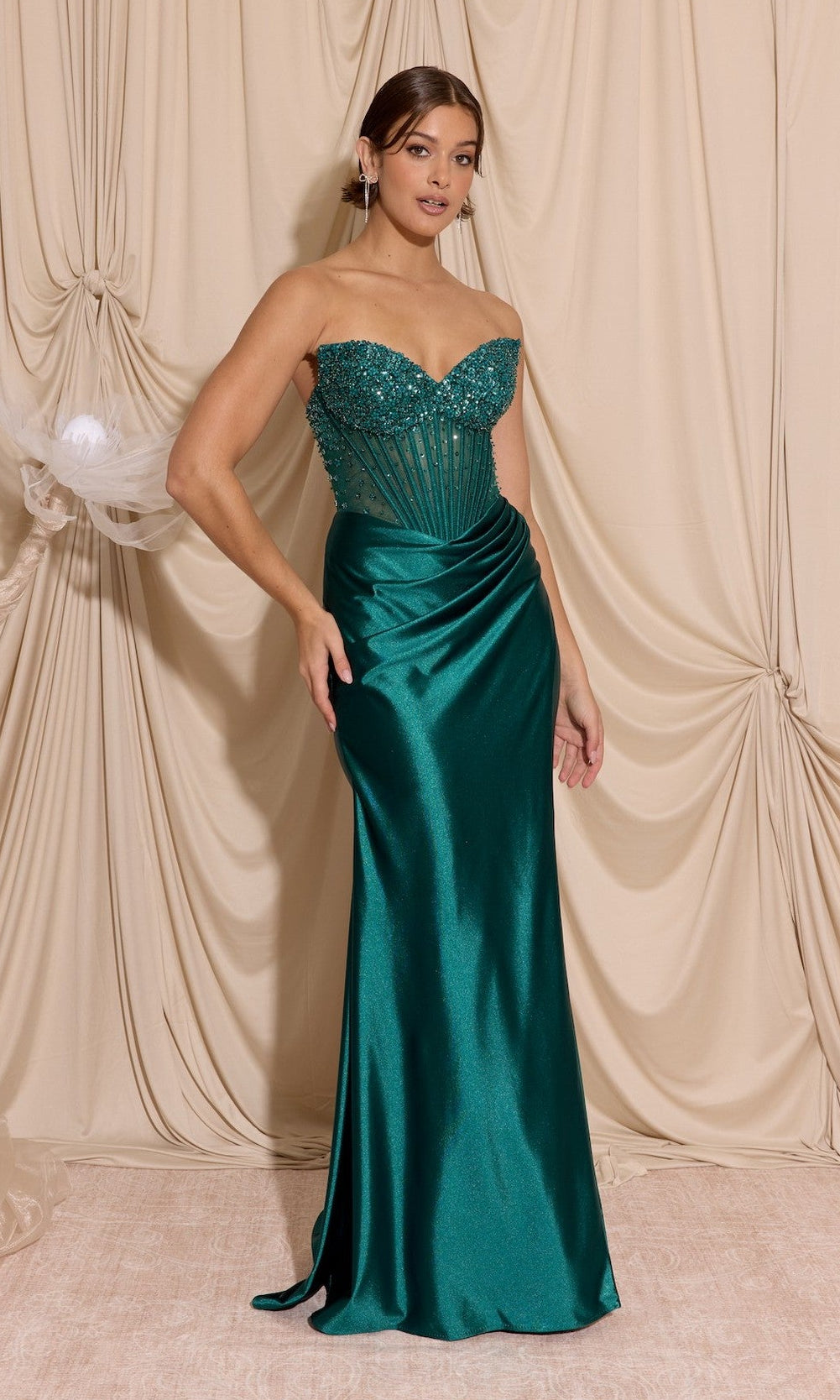 A woman in the Strapless Tight Satin Prom Dress: Eureka EK183 in emerald green, with a beaded bodice and corset detailing, stands before beige draped curtains—an ideal choice for formal events.