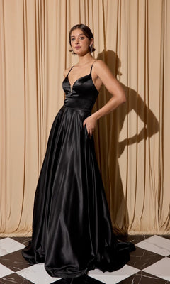 A woman stands before beige curtains wearing the Eureka EK190 Long Prom Dress in black, featuring thin straps, a fitted bodice, and an elegant floor-length A-line skirt.