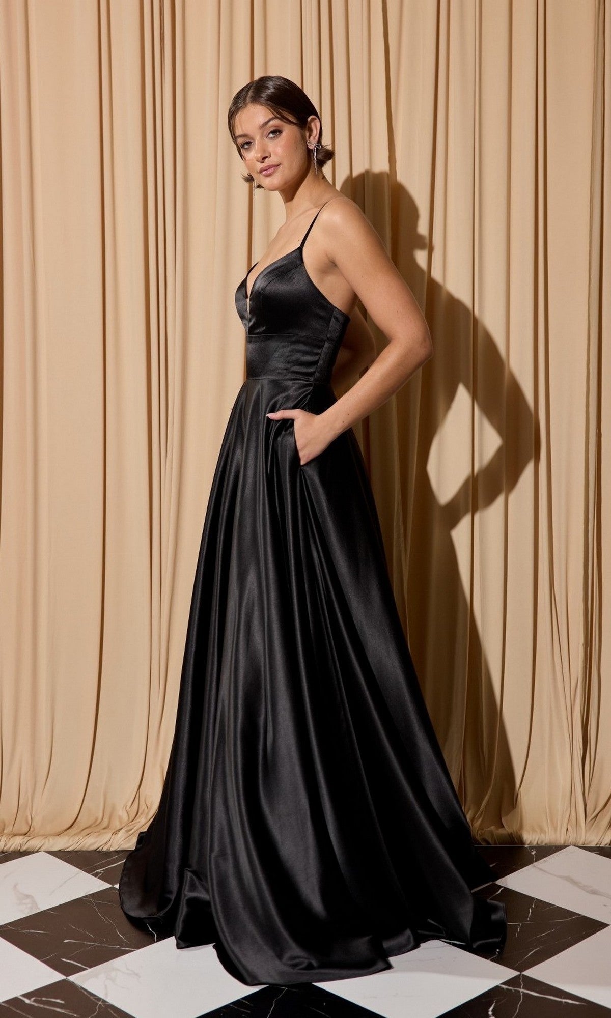 A woman wearing the Eureka EK190 long prom dress, a sleeveless black satin gown with pockets and an A-line skirt, stands on a black and white tiled floor in front of beige curtains.