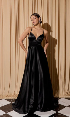 A woman poses in front of beige curtains on a black-and-white tiled floor wearing the Eureka EK190 long prom dress, featuring black satin fabric, a deep V neckline, open back, A-line skirt, and pockets.