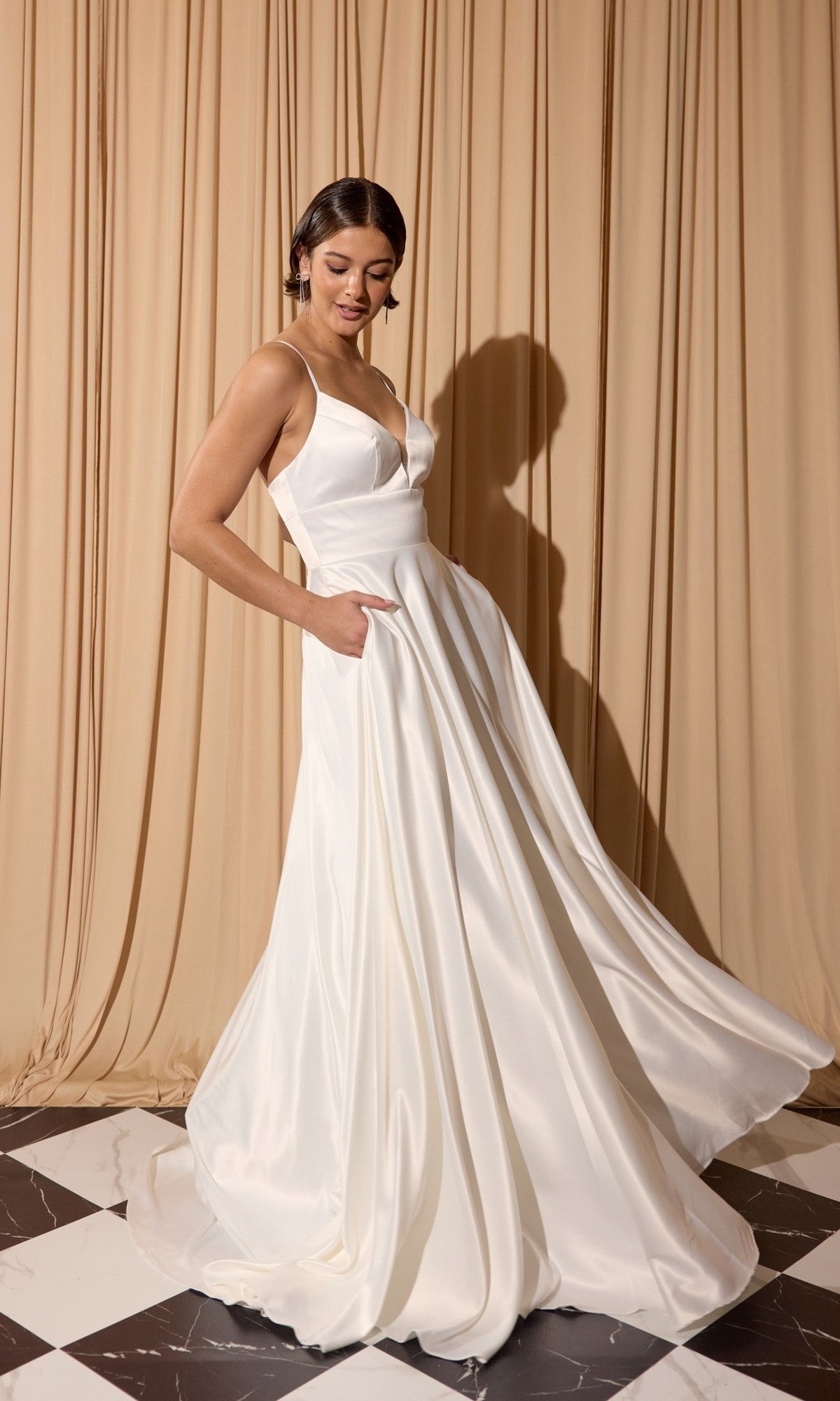 A woman in the Eureka EK190 Long Prom Dress in Off White, featuring an a-line skirt and pockets, stands on a black and white tile floor, posing before beige curtains.