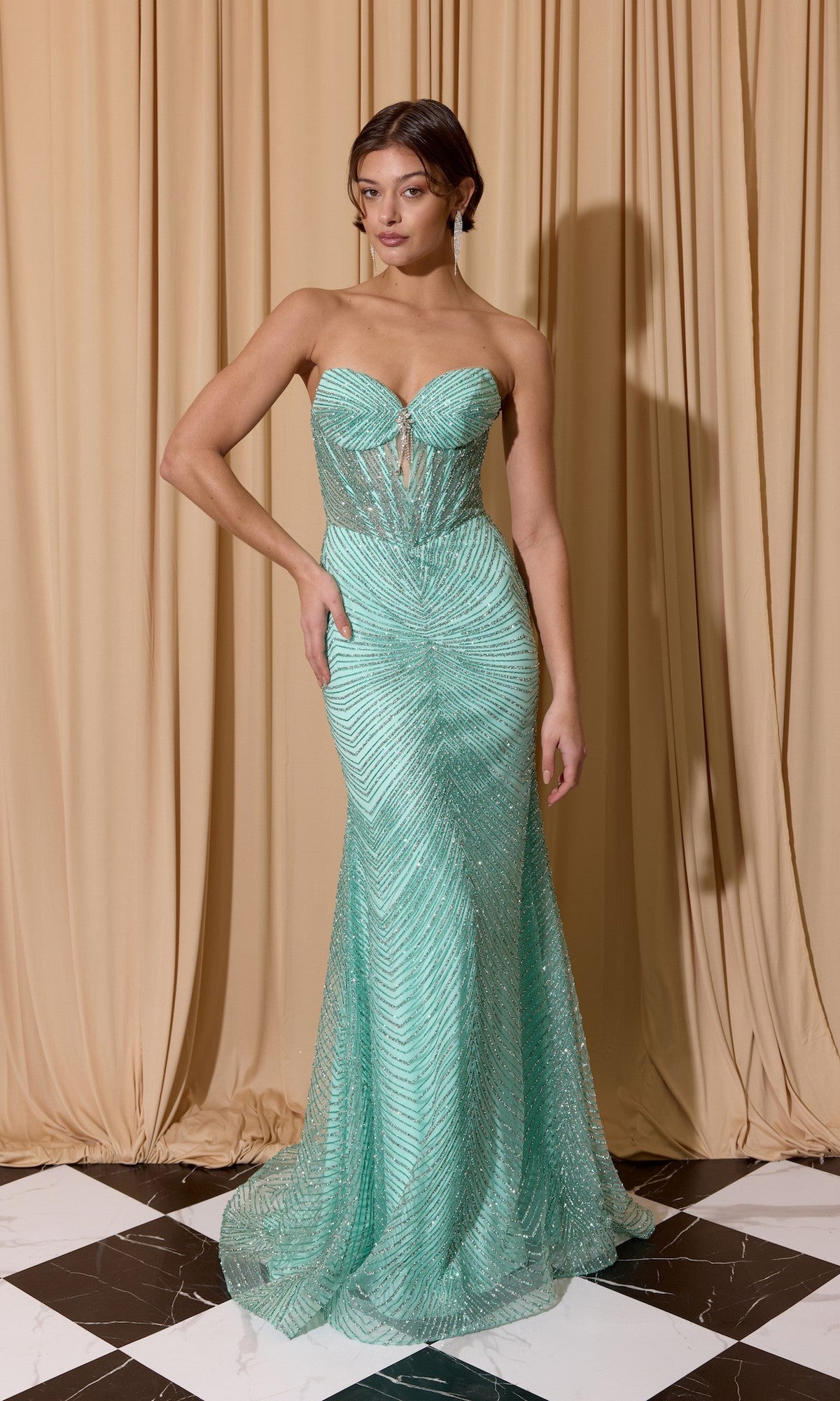A woman stands before beige curtains on black and white tiles, wearing the Eureka EK195—a mint green, strapless prom dress with a fitted corset bodice, sheer patterned design, and floor-length hem.