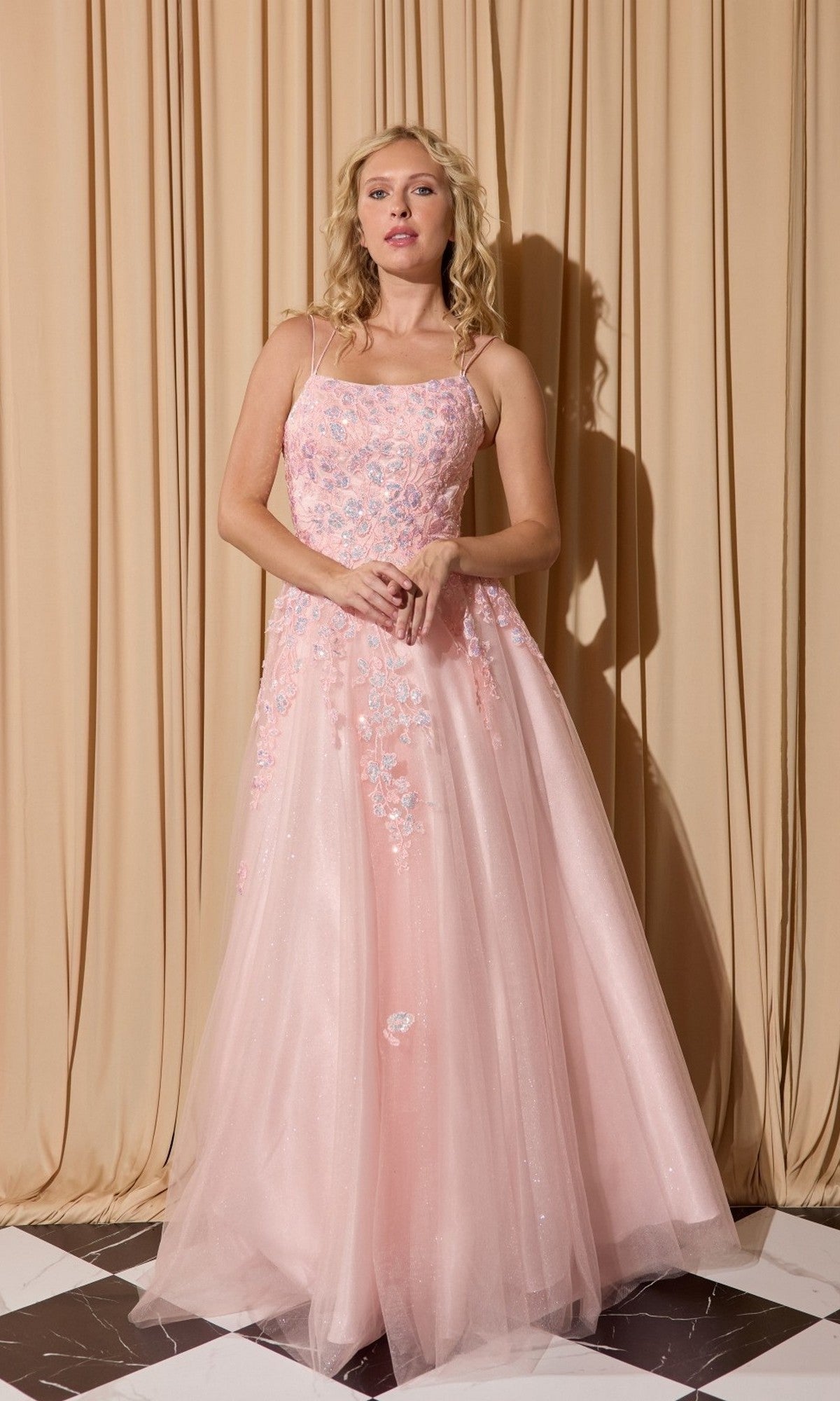 A woman stands before beige curtains wearing the Eureka EK197 long prom dress, a floor-length, light pink glitter tulle gown with delicate floral embroidery and thin straps.