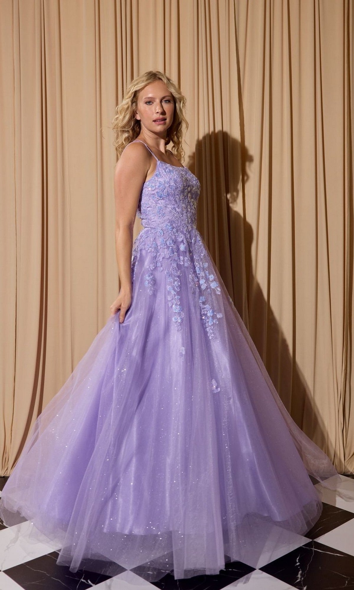 A woman stands on a black and white checkered floor wearing the Eureka EK197 long lilac prom dress with floral appliqués, in front of beige curtains.