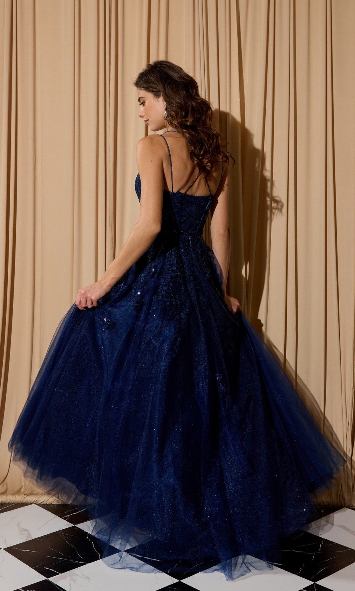 A woman wearing the Eureka EK197 long prom dress in dark blue glitter tulle stands on a black-and-white tiled floor, facing away, with beige curtains behind her.