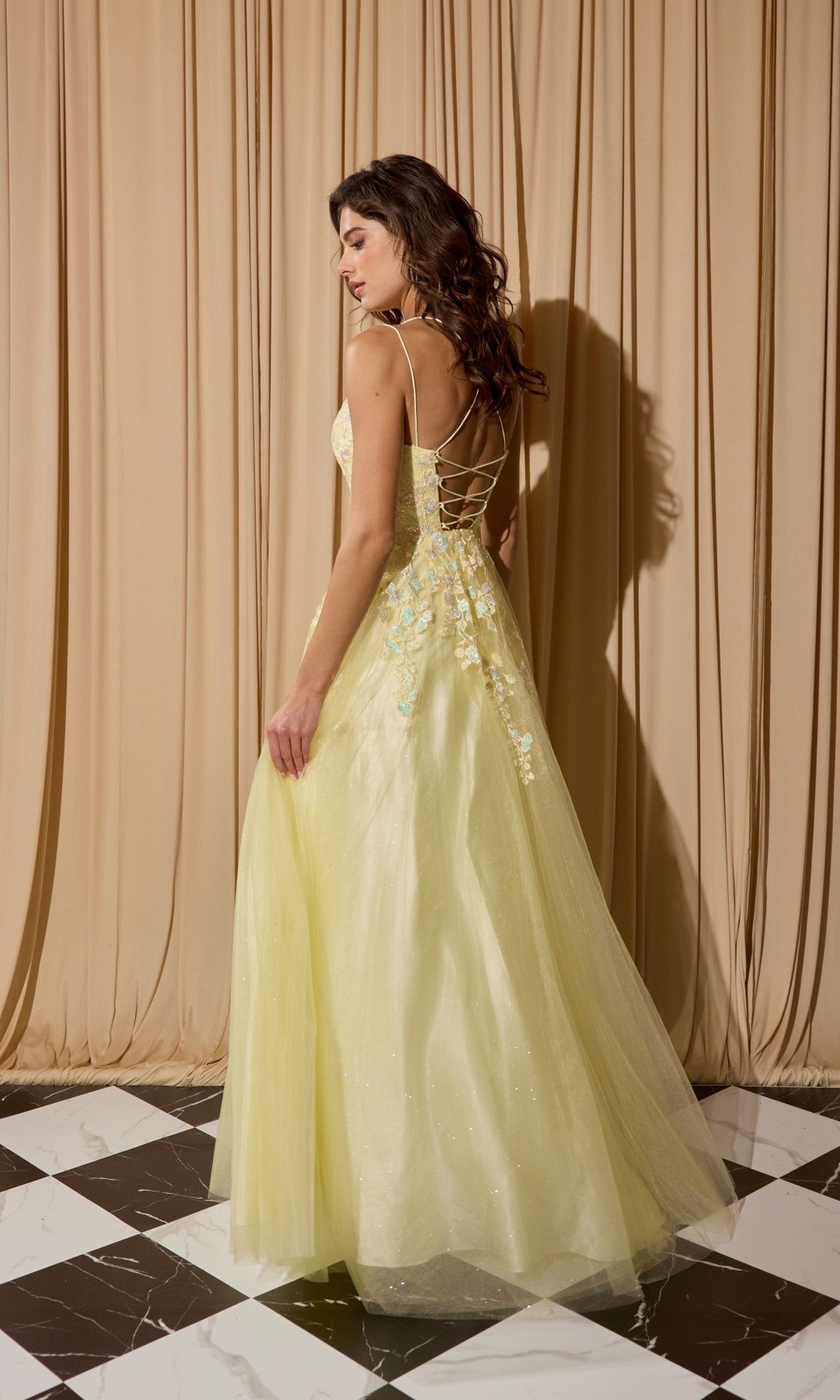 A woman wearing the Eureka EK197 long prom dress with yellow lace appliques, a lace-up back, and floral details stands on a black-and-white checkered floor before beige curtains.