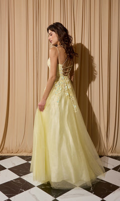 A woman wearing the Eureka EK197 long prom dress with yellow lace appliques, a lace-up back, and floral details stands on a black-and-white checkered floor before beige curtains.
