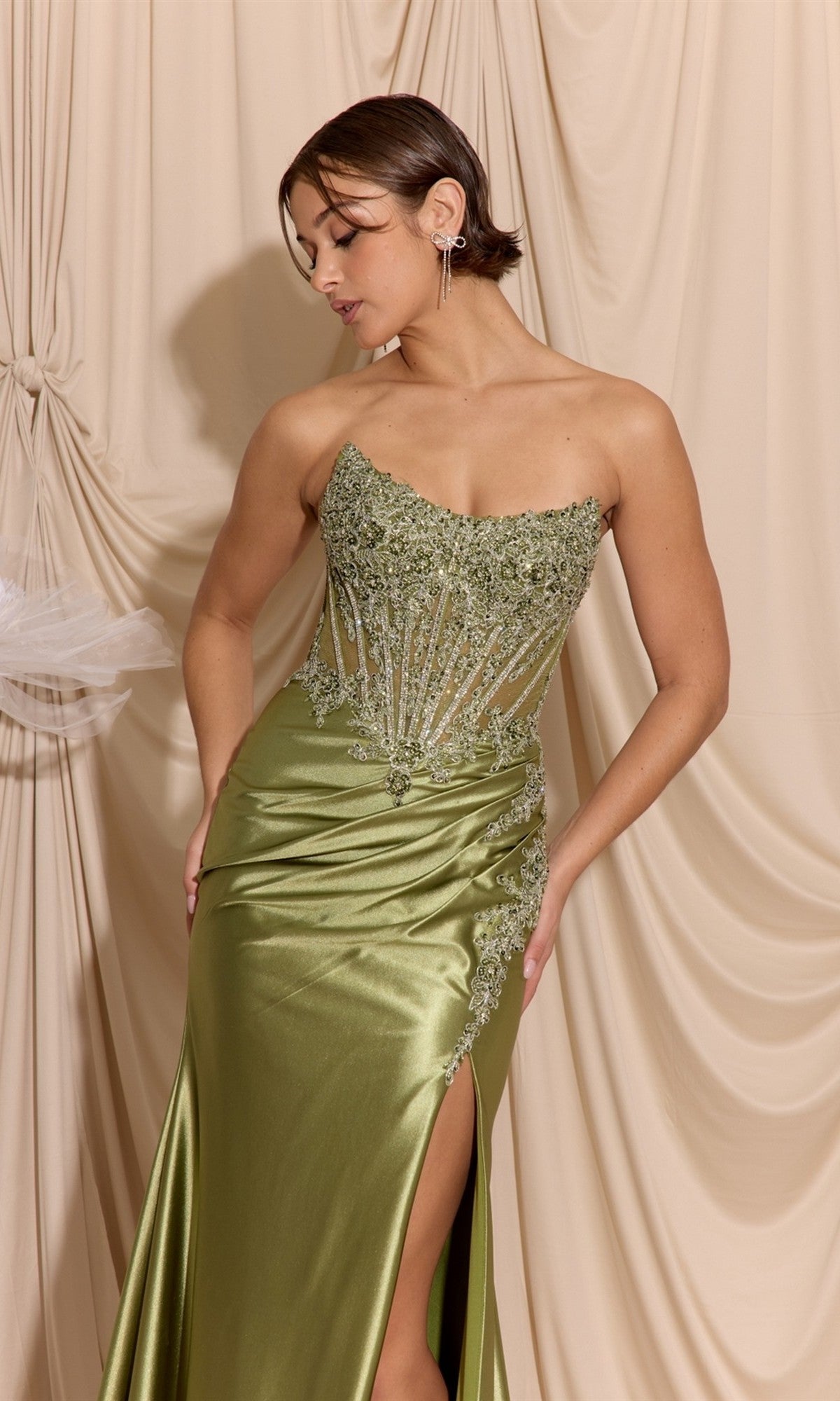 A woman wearing the Eureka EK202 long prom dress in olive green, featuring a strapless beaded embroidered bodice and satin sheath skirt, stands against draped beige fabric, looking down with one hand on her hip.