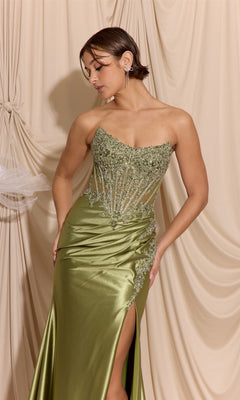 A woman wearing the Eureka EK202 long prom dress in olive green, featuring a strapless beaded embroidered bodice and satin sheath skirt, stands against draped beige fabric, looking down with one hand on her hip.