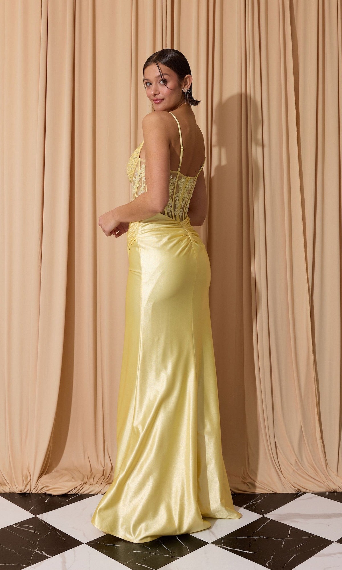 A woman in the Eureka EK203 long prom dress, featuring a yellow satin sleeveless design with a lace corset bodice and sheath skirt, poses on a black and white floor with her back slightly turned before beige draped curtains.