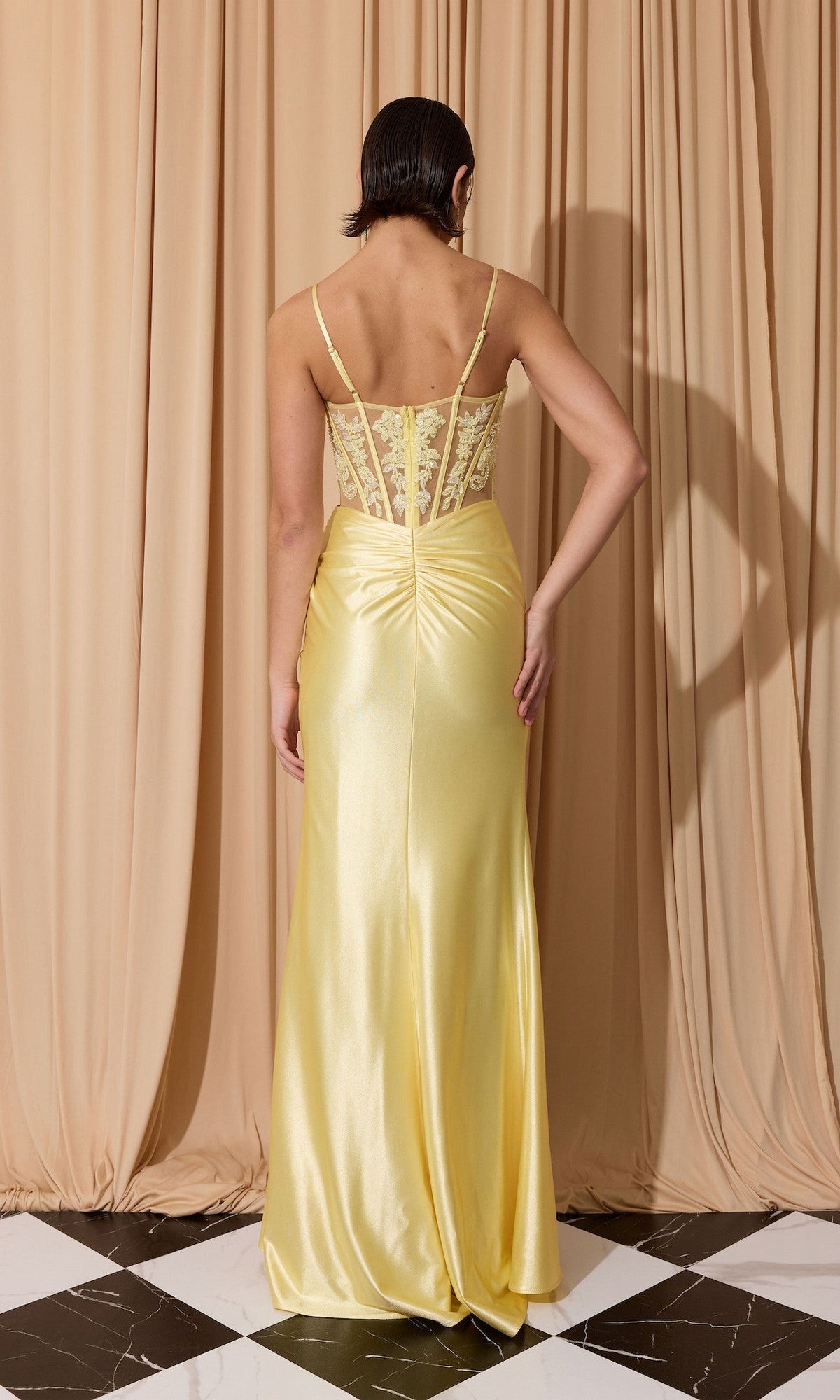 A person faces away from the camera, wearing the Eureka EK203 Long Prom Dress—a yellow satin gown with a sheath skirt and sheer, embroidered corset bodice—standing before beige curtains on a black and white tiled floor.