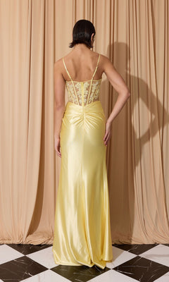 A person faces away from the camera, wearing the Eureka EK203 Long Prom Dress—a yellow satin gown with a sheath skirt and sheer, embroidered corset bodice—standing before beige curtains on a black and white tiled floor.