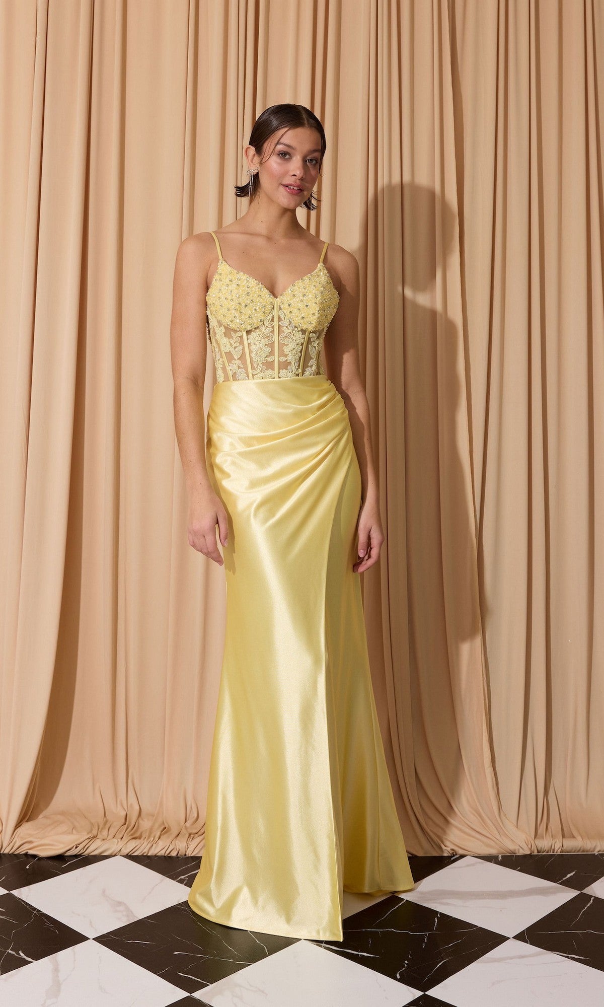 A woman stands before beige curtains wearing the Eureka EK203, a floor-length yellow satin prom dress with a fitted corset bodice, delicate embellishments, and spaghetti straps.