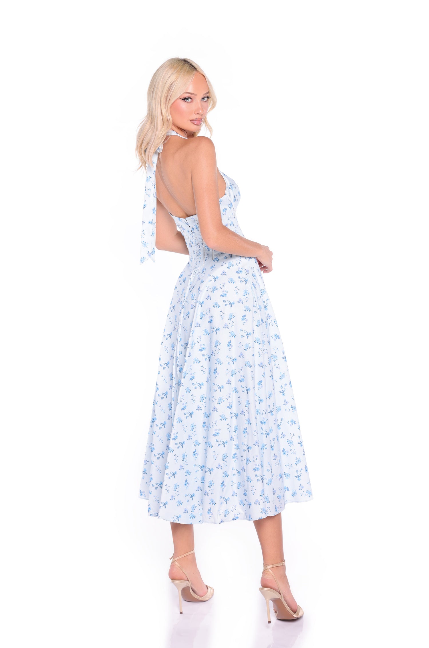 A woman models the Light Blue Floral Halter Summer Midi Dress: Eliza, standing in high heels and facing slightly backward against a white background.