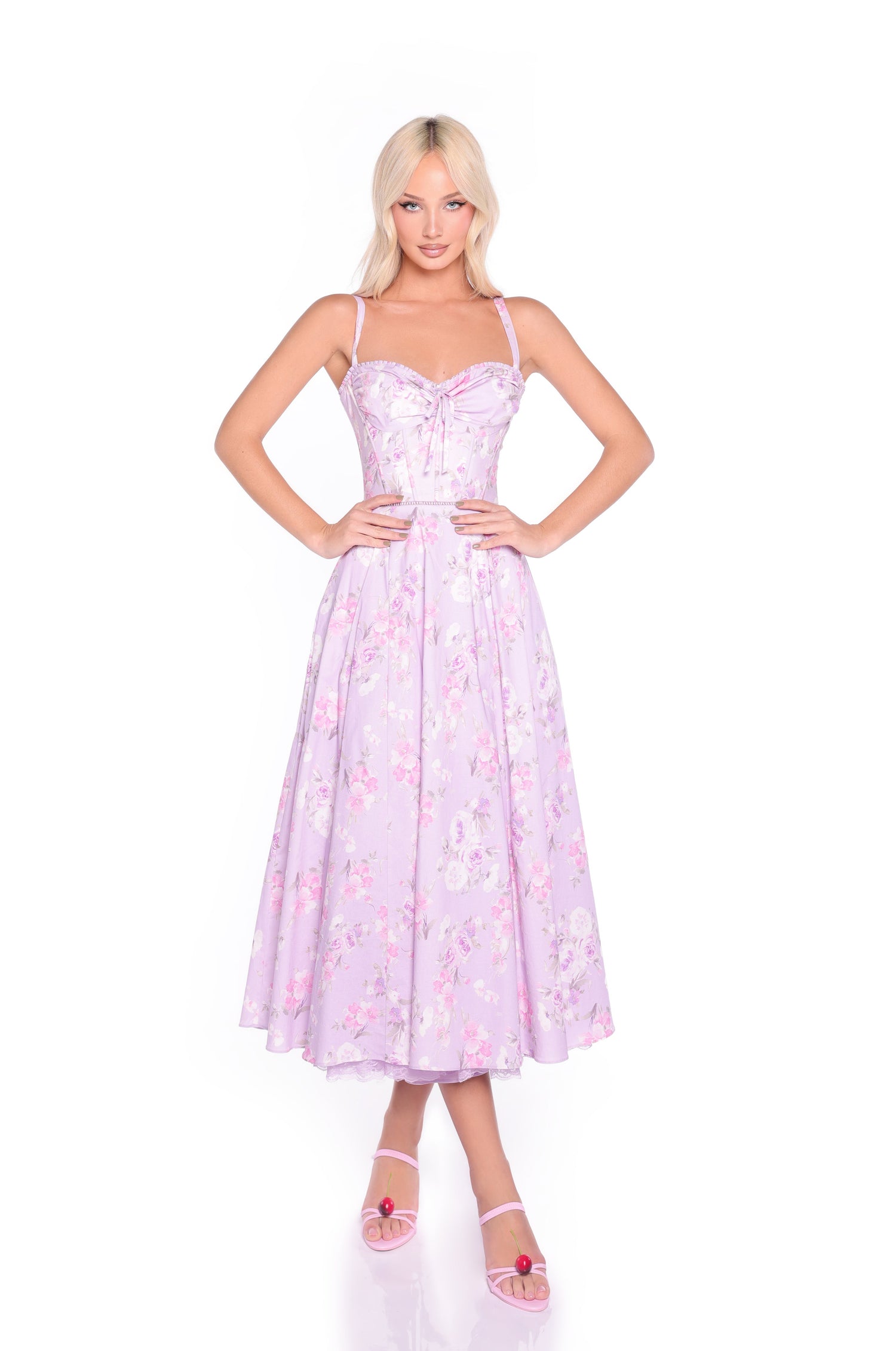 A woman faces forward in the Elle Lilac Floral Print Midi Summer Dress, featuring a corset bodice and flared skirt, paired with light pink open-toe heels.