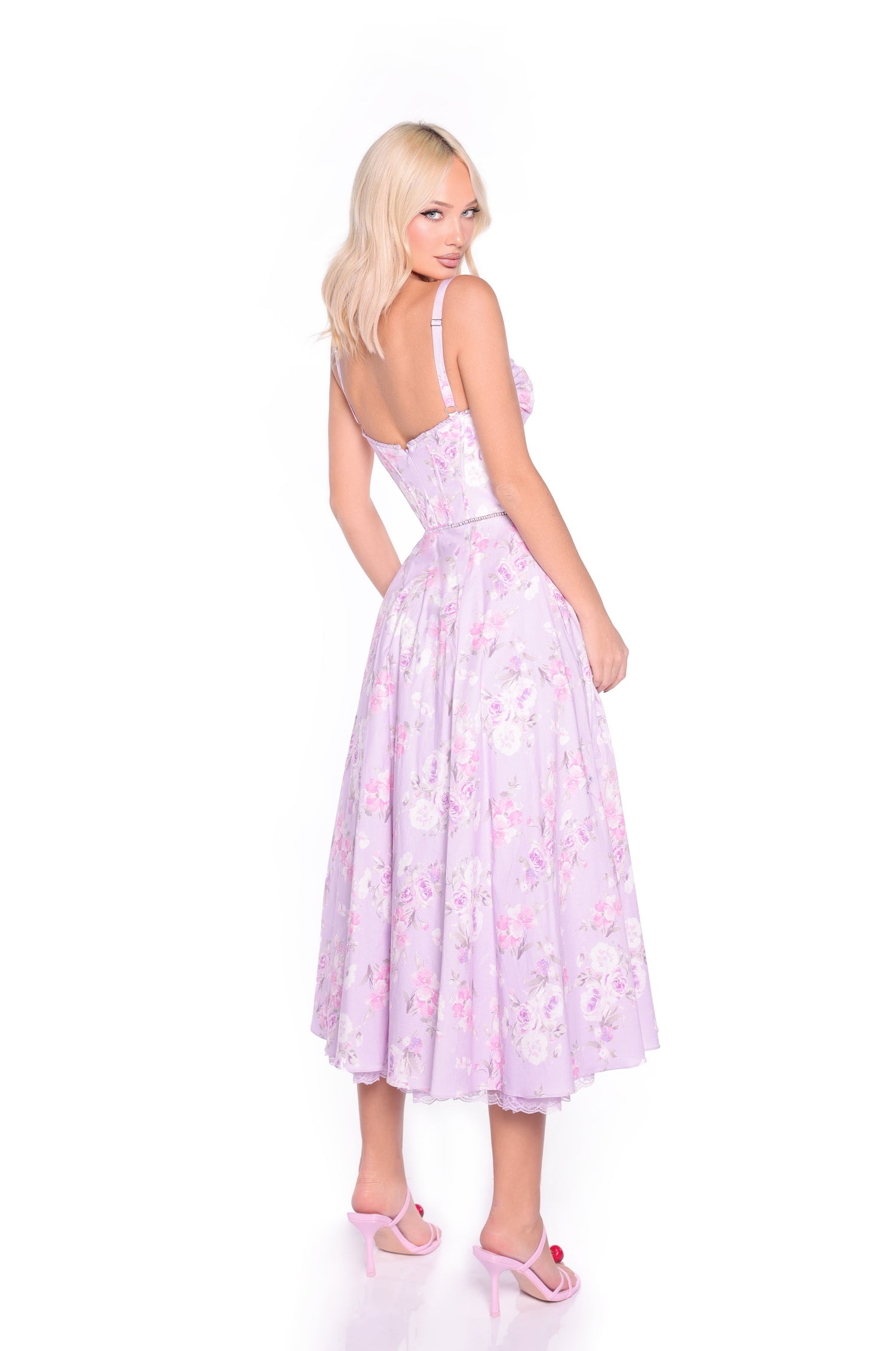 A blonde woman stands against a white background, wearing the Elle Lilac Floral Print Midi Summer Dress with a corset bodice and pink heels, looking over her shoulder.