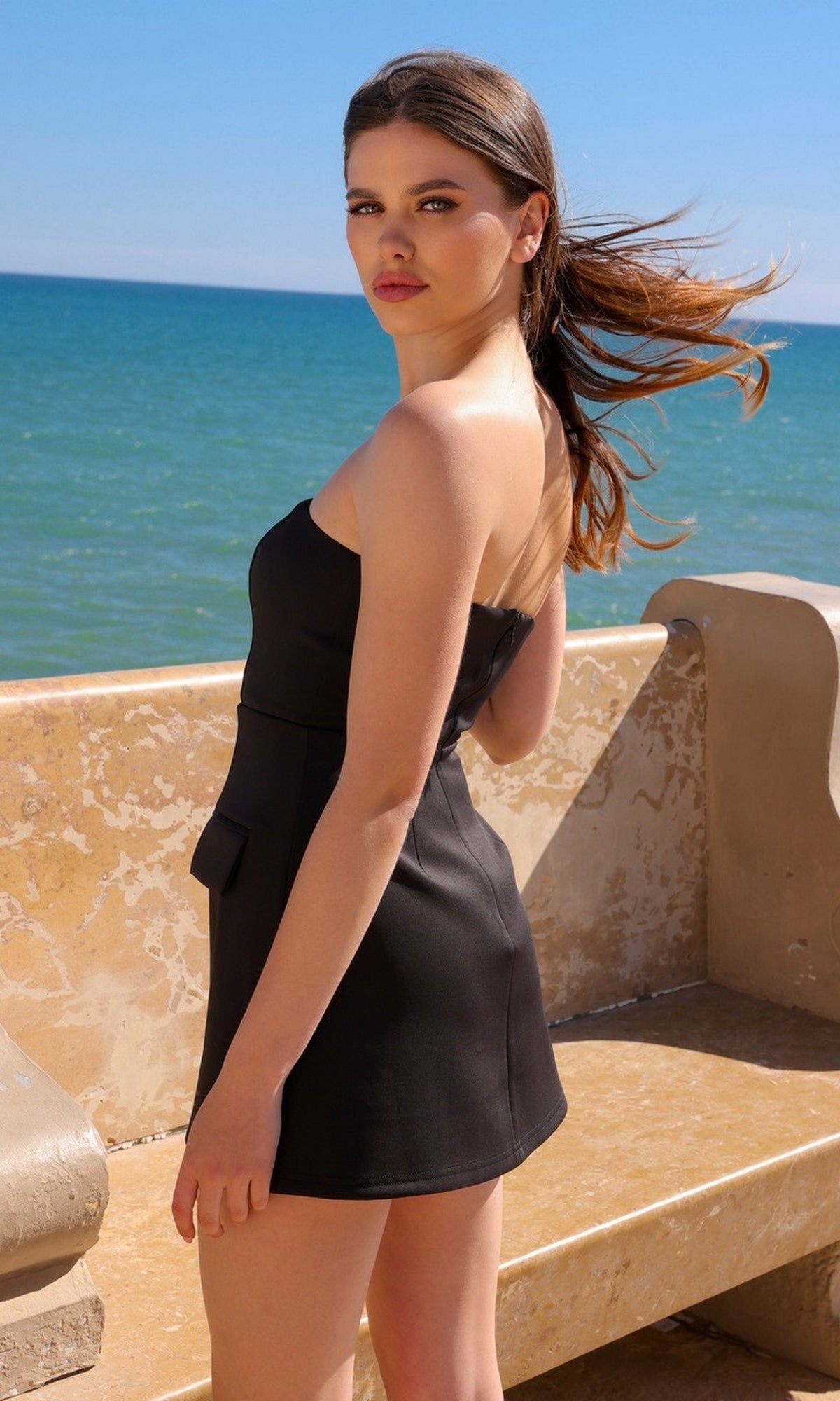 A woman wearing the Velvi Strapless Mini Graduation Dress Ellis stands by a stone bench near the ocean, looking over her shoulder. The sky is clear and her hair blows in the wind.
