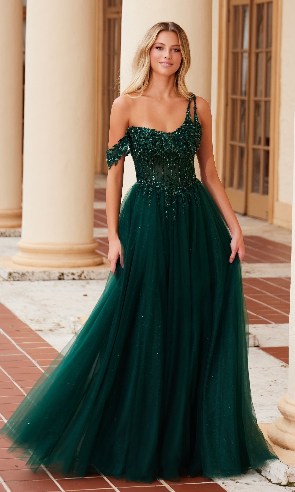 A woman in the Nox Anabel F1567 Asymmetrical Glitter Prom Dress poses on a tiled patio with cream columns and glass doors in the background.
