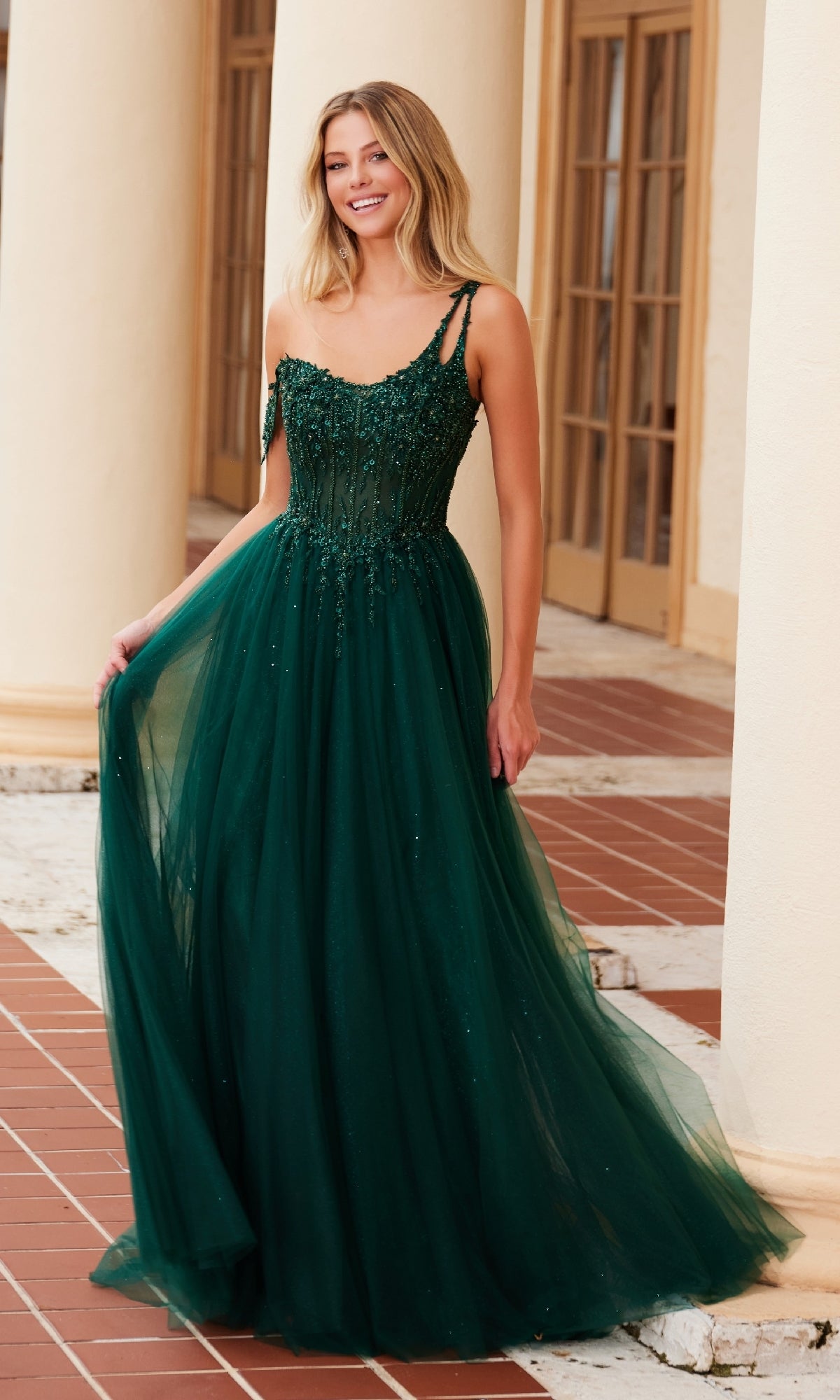 A woman smiles as she stands on a tiled floor near light columns, holding one side of her Asymmetrical Glitter Prom Dress: Nox Anabel F1567, featuring dark green beaded embroidery and a tulle skirt.