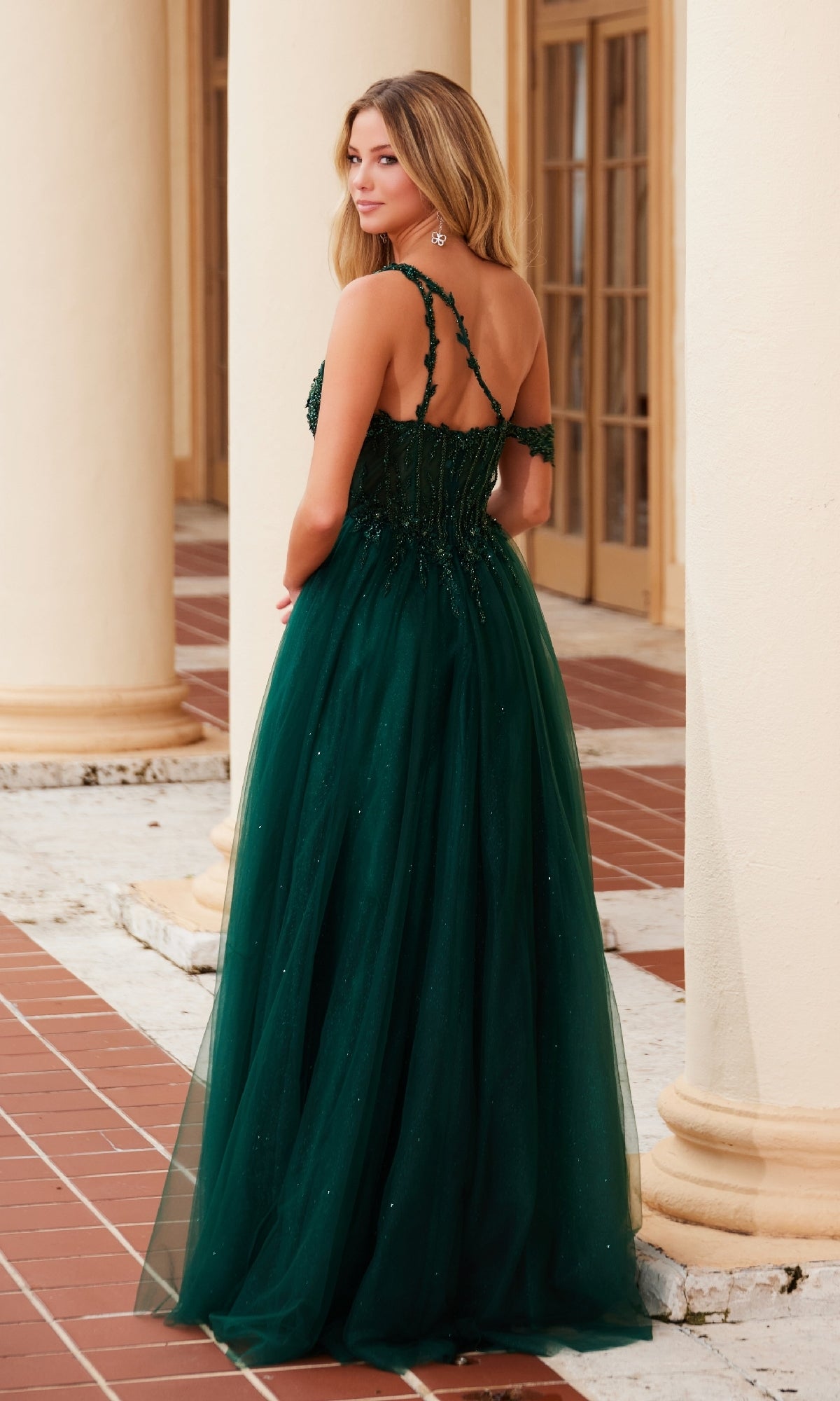 A woman in the Asymmetrical Glitter Prom Dress Nox Anabel F1567 stands outdoors near cream-colored columns, elegantly looking over her shoulder.