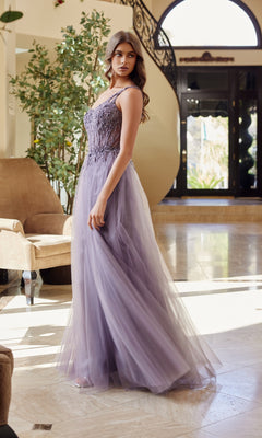 A woman stands indoors on a marble floor near a chair and staircase, wearing the Nox Anabel F1567 Asymmetrical Glitter Prom Dress featuring long lavender tulle and thin straps.
