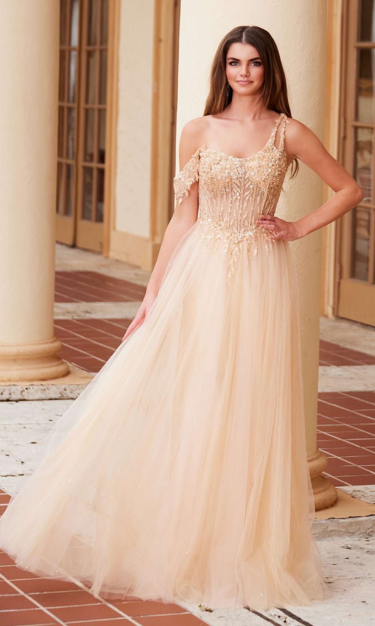 A woman poses outdoors with one hand on her hip, wearing the Nox Anabel F1567 Asymmetrical Glitter Prom Dress— a floor-length beige gown featuring sequin details and a glitter tulle skirt, in front of columns and glass doors.