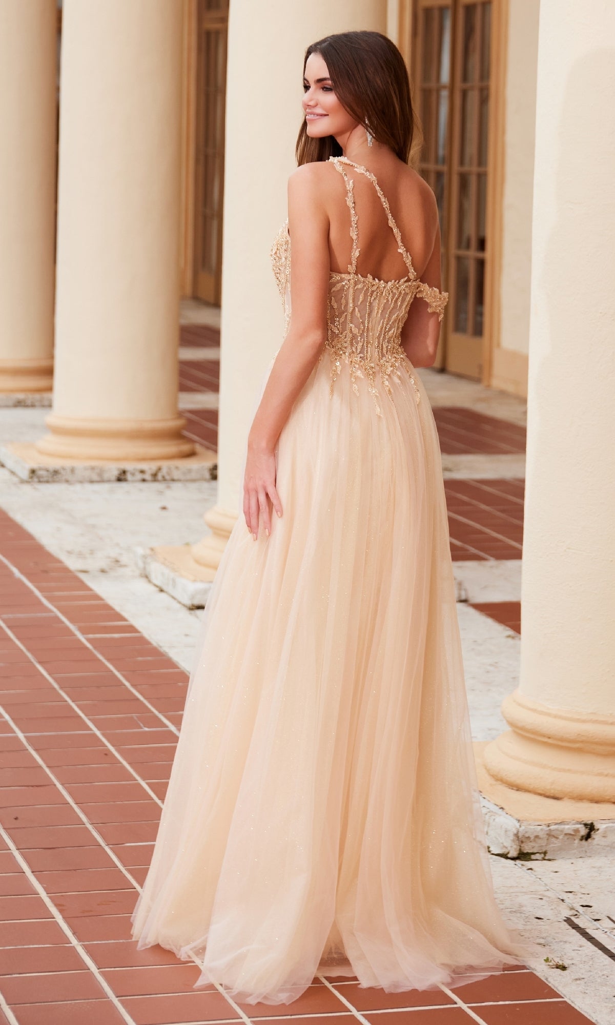 A woman in the Nox Anabel F1567 Asymmetrical Glitter Prom Dress, featuring a light beige, floor-length skirt and sheer straps, stands on a tiled walkway with her back to the camera.
