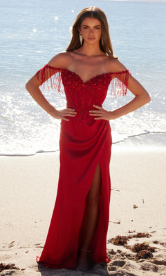 A woman stands on a sandy beach in the Beaded-Bodice Long Prom Dress: Nox Anabel F1571—a red, off-the-shoulder gown with a beaded bodice, floor-length skirt, fringe details, and thigh-high slit—while ocean water shimmers behind her.