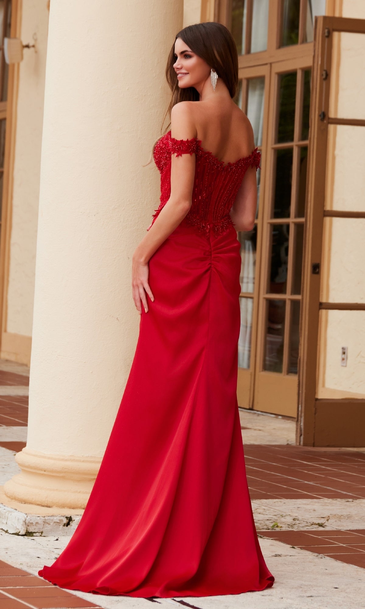 A woman wearing the Beaded-Corset Long Prom Dress: Nox Anabel F1572 in red stands outside by a cream-colored column, facing away from the camera.
