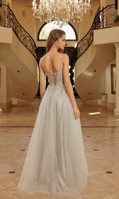 A woman wears the Nox Anabel F1726 long prom dress, featuring a sleeveless beaded corset bodice and lace-up back, as she stands in an elegant foyer with marble floors, curved staircases, and a sparkling chandelier.