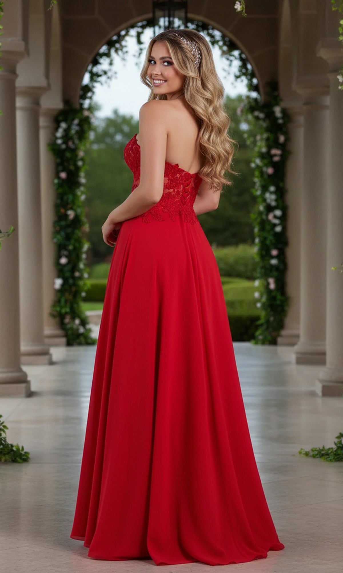 A woman wearing the Long Red Sheer-Bodice Prom Dress by PromGirl stands in an outdoor colonnade, facing away but glancing back over her shoulder, with lush greenery in the background.