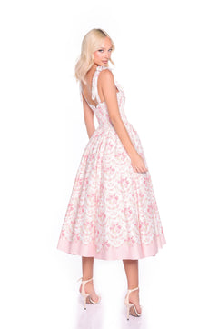 A woman poses in the Pink Floral Casual Midi Dress: Florence, a sleeveless A-line with ruffled straps, paired with pink heels. Perfect for casual weddings or sunny days, she stands slightly turned against a white background.