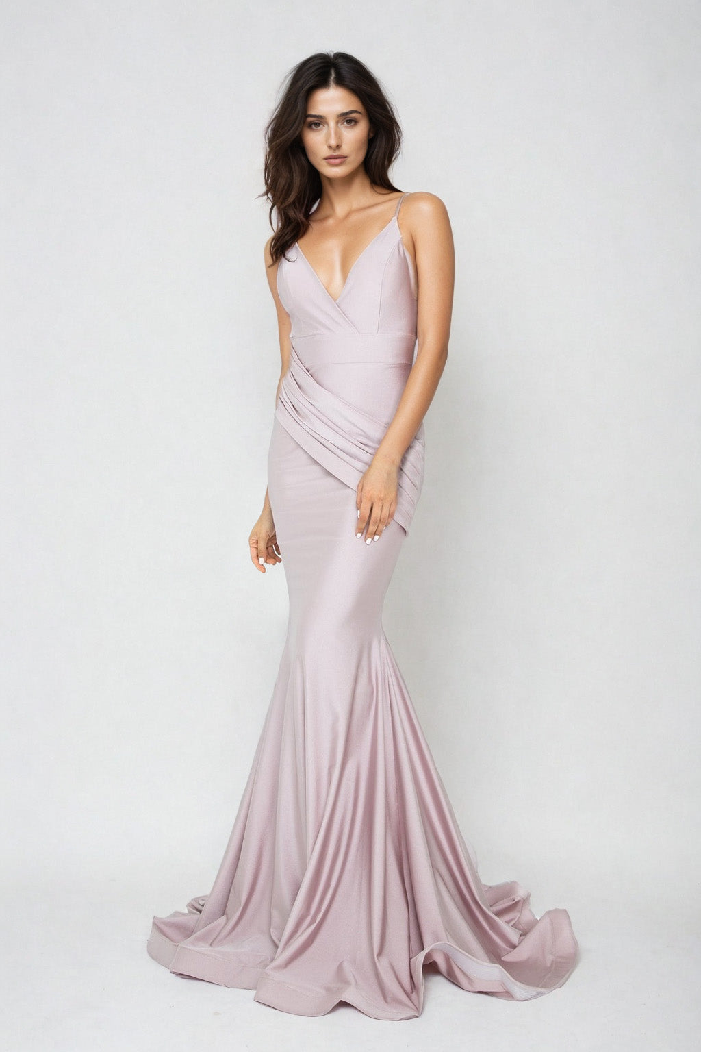A woman stands against a plain background wearing the Rushed-Sash Long Mermaid Prom Dress: Atria 6200H, a fitted, light mauve, floor-length gown with a deep V-neckline and thin straps.