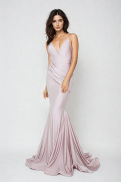 A woman stands against a plain background wearing the Rushed-Sash Long Mermaid Prom Dress: Atria 6200H, a fitted, light mauve, floor-length gown with a deep V-neckline and thin straps.
