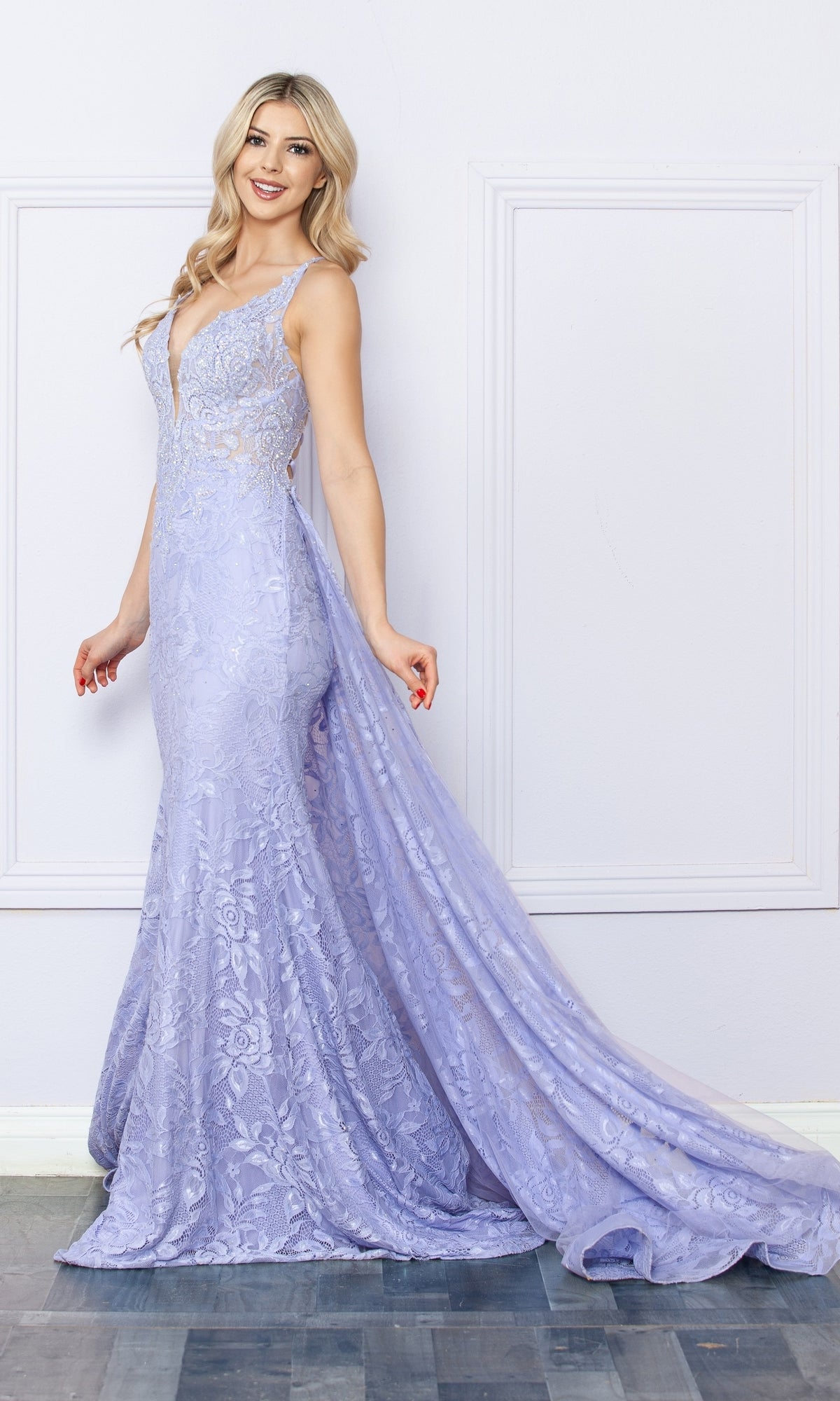 A woman in the Open-Back Long Lace Prom Dress with Train G1353, a floor-length light purple floral lace mermaid gown, stands against a white wall looking over her shoulder and smiling.