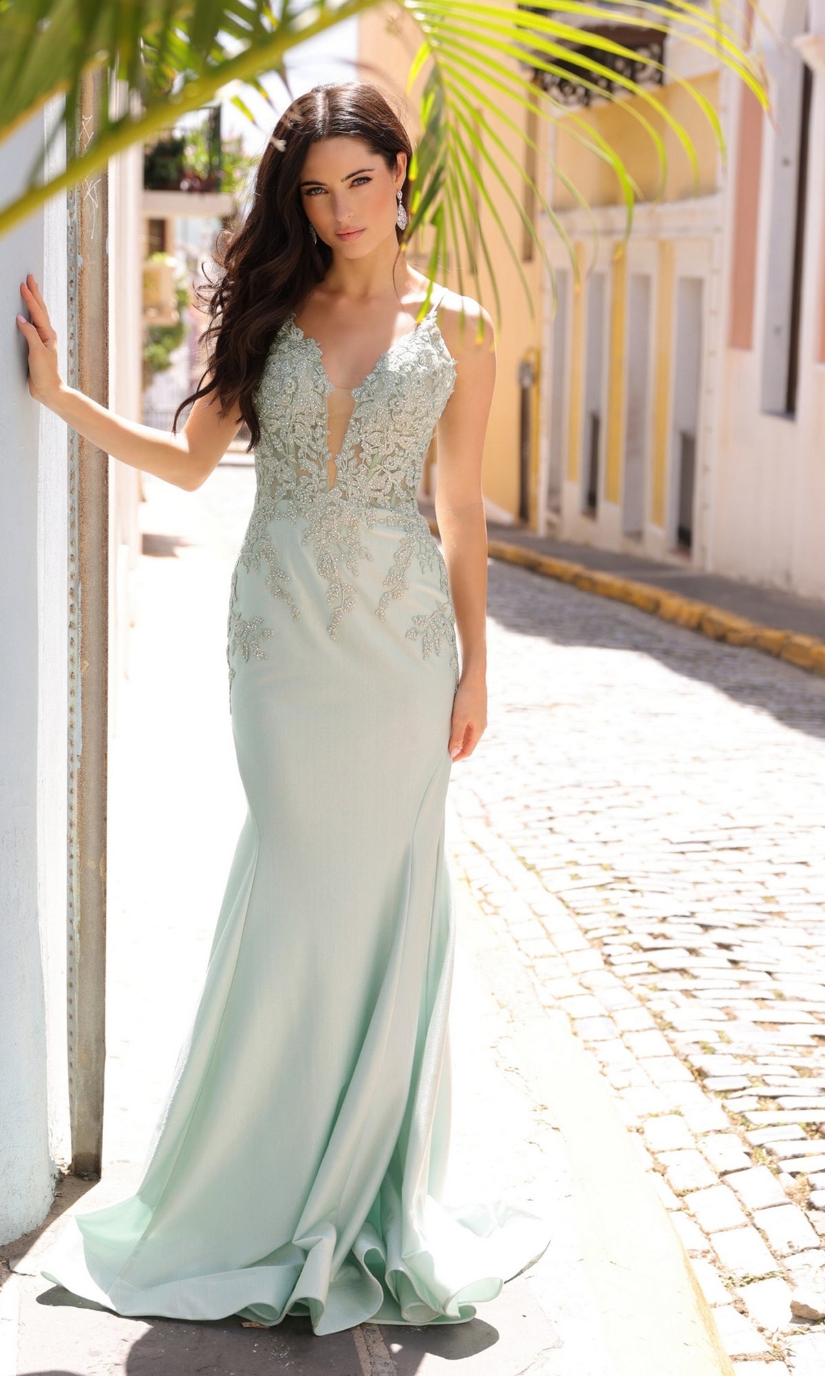 A woman in the Lace-Up Embroidered Bodice Long Prom Dress G1364, a light blue sleeveless floor-length gown with beaded lace embroidery, stands on a sunny cobblestone street, leaning casually against a wall.